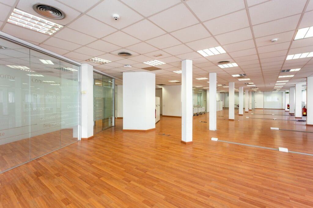 Large Empty Space With Ceiling Tiles, Fluorescent Lights, Light Brown Laminate Flooring And White Painted Columns. Large Office Space For Rent Just Renovated And Unfurnished